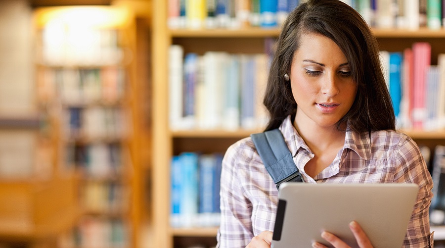 Garota estudante bonita segurando e olhando para um tablet dentro de uma biblioteca