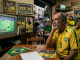 Foto mostrando o interior de um boteco tradicional brasileiro chamado 'Boteco do Jorge'. Um homem idoso com barba e camisa amarela da Seleção Brasileira com o número 9 apoia o rosto na mão, com uma expressão triste e pensativa. À sua frente, na mesa de madeira, há um caderno aberto com anotações sobre futebol, uma foto antiga da conquista de 2002 e uma pequena bandeira do Brasil. Ao fundo, uma televisão antiga exibe o jogador Neymar andando de cabeça baixa na Copa de 2022, enquanto outros torcedores no bar expressam desapontamento. Nas paredes há quadros com fotos históricas de Pelé e Ronaldo Fenômeno erguendo a taça.